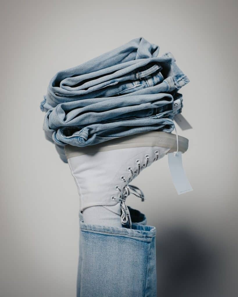Eine Person, die eine hellblaue Jeans und einen weißen High-Top-Sneaker trägt, hält einen Stapel gefalteter blauer Jeans auf der Sohle ihres erhobenen Fußes vor einem schlichten hellen Hintergrund. Eine Person, die eine hellblaue Jeans und einen weißen High-Top-Sneaker trägt, hält einen Stapel gefalteter blauer Jeans auf der Sohle ihres erhobenen Fußes vor einem schlichten hellen Hintergrund.