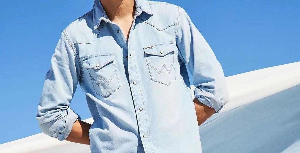 Eine Person, die ein hellblaues Jeanshemd mit aufgekrempelten Ärmeln trägt, steht im Freien vor einem strahlend blauen Himmel und weißem Hintergrund. Ihr Kopf ist auf dem Bild nicht zu sehen.