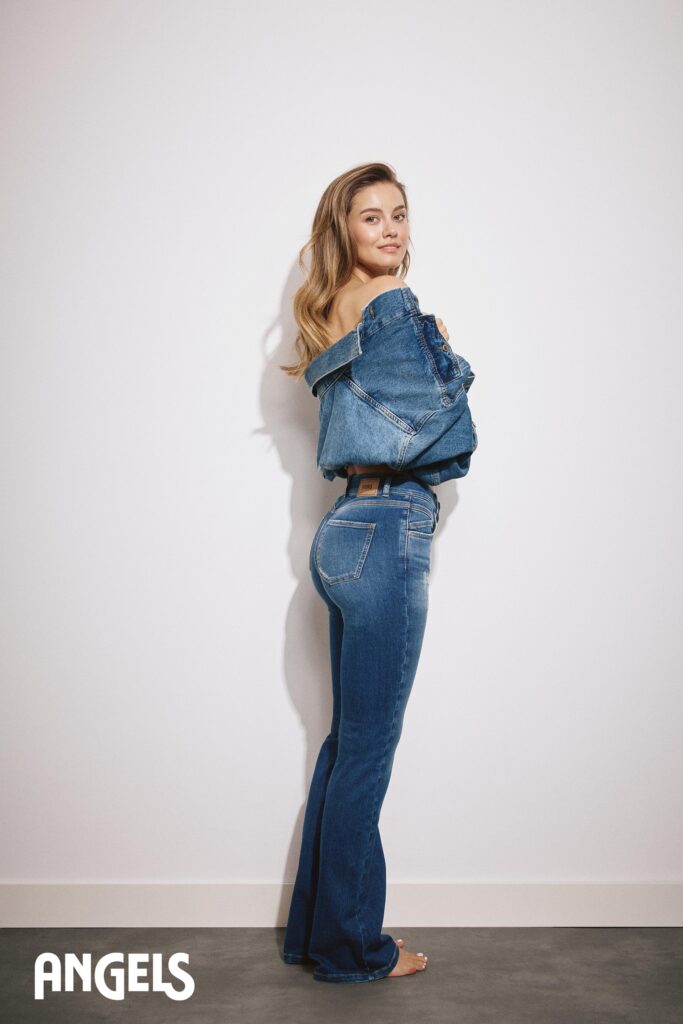 Eine Frau in Jeansjacke und Jeans steht barfuß vor einer weißen Wand, schaut über ihre Schulter und lächelt. In der unteren linken Ecke ist das Wort ANGELS aufgedruckt.