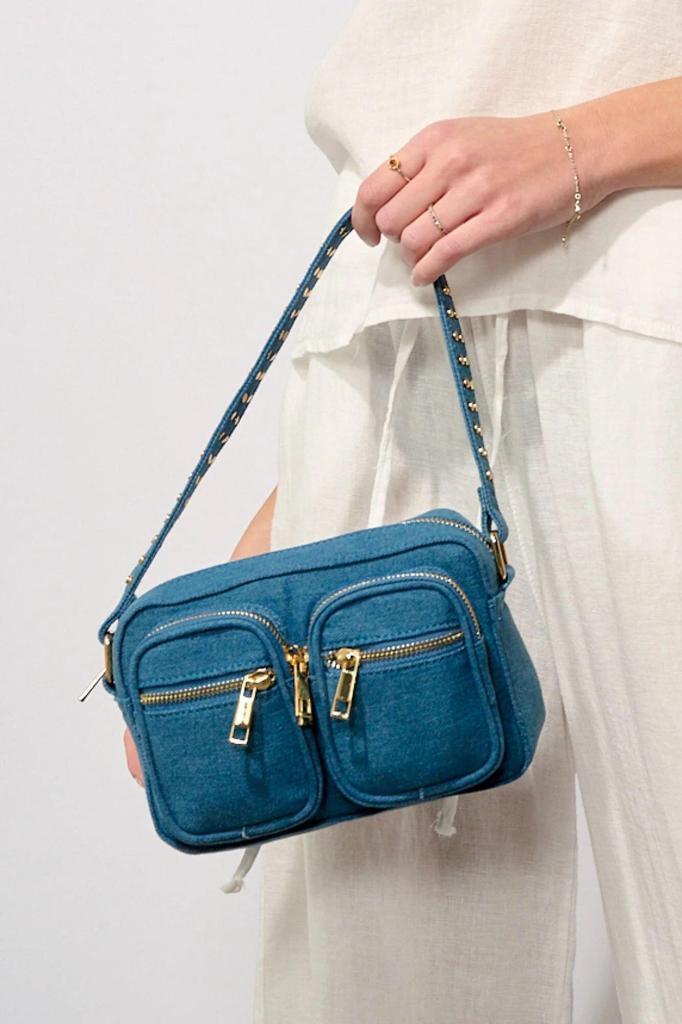 eine person in weißem outfit hält eine blaue handtasche mit goldenen reißverschlüssen und beschlägen. die tasche hat zwei vordertaschen mit reißverschluss und einen schulterriemen mit nieten. Eine weiß gekleidete Person hält eine blaugrüne Handtasche mit goldenen Reißverschlüssen und zwei Vordertaschen sowie einem kurzen, mit goldenen Nieten verzierten Henkel.