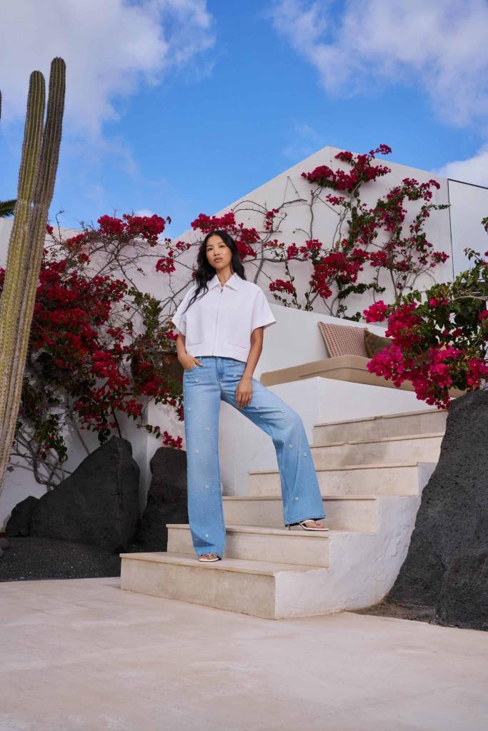 Eine Frau steht auf weißen Steinstufen im Freien und trägt ein weißes kurzärmeliges Hemd und hellblaue Jeans. Rote Blumen und grüne Kakteen umgeben sie, im Hintergrund ein weißes Gebäude und blauer Himmel. Eine Frau steht auf weißen Steinstufen im Freien und trägt ein weißes kurzärmeliges Hemd und hellblaue Jeans. Rote Blumen und grüne Kakteen umgeben sie, im Hintergrund ein weißes Gebäude und blauer Himmel.