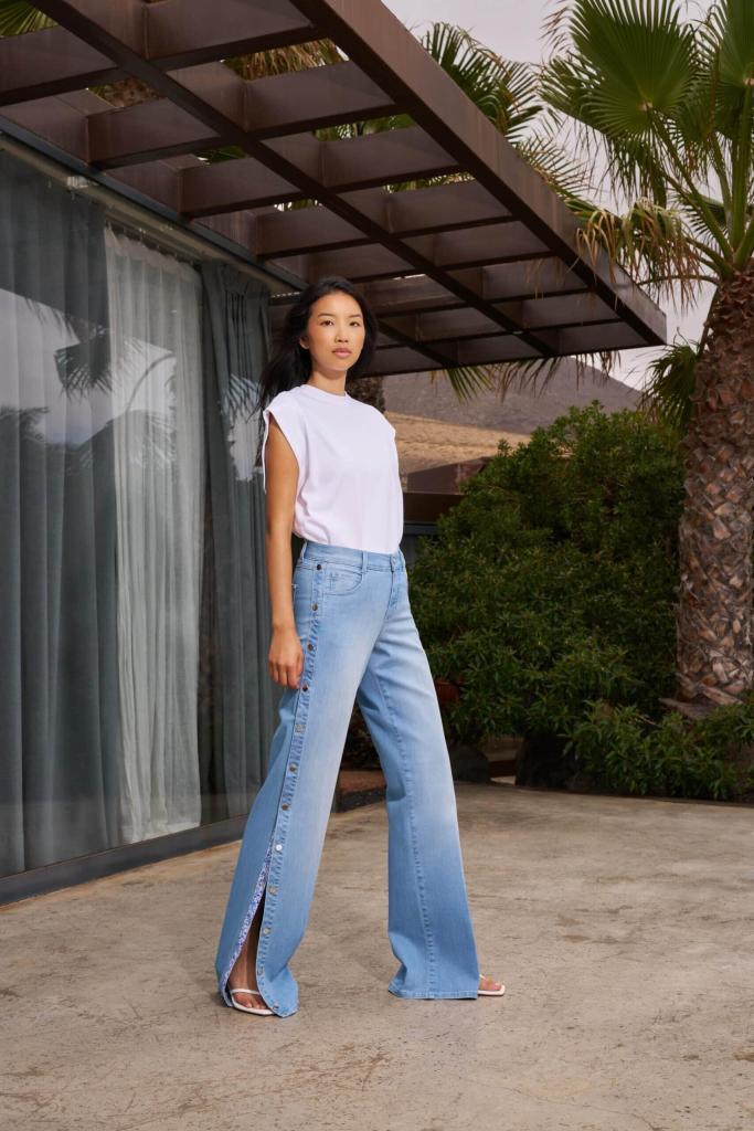 Eine Frau steht auf einer Terrasse und trägt ein weißes, ärmelloses Oberteil und hellblaue, weit geschnittene Jeans mit hohen Seitenschlitzen. Sie posiert selbstbewusst vor einem modernen Gebäude mit Glaswänden und umgeben von Grün. Eine Frau steht auf einer Terrasse und trägt ein weißes, ärmelloses Oberteil und hellblaue, weit geschnittene Jeans mit hohen Seitenschlitzen. Sie posiert selbstbewusst vor einem modernen Gebäude mit Glaswänden und umgeben von Grün.