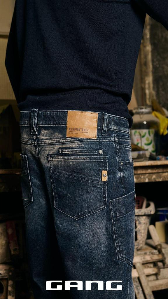 Eine Person, die dunkelblaue Jeans und einen schwarzen Pullover trägt, steht mit dem Rücken zur Kamera. Der Lederaufnäher auf der Jeans zeigt die Marke GANG. Im Hintergrund sind unscharfe Werkstattgegenstände zu sehen.