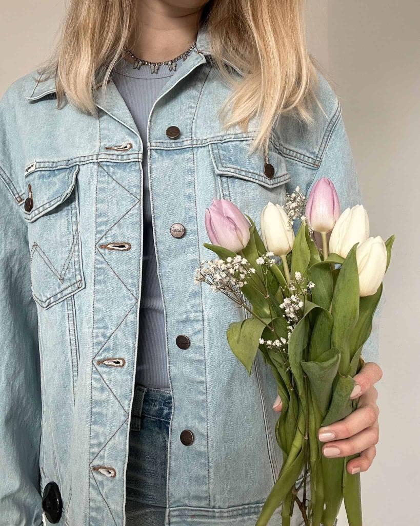 Eine Person mit langem blondem Haar, die eine hellblaue Jeansjacke trägt und einen Strauß weißer und hellrosa Tulpen mit Grünzeug und Babyatemblumen in der Hand hält. Ihr Gesicht ist teilweise außerhalb des Rahmens. Frau in hellblauer Oversize-Jeansjacke haelt rosa und weisse Tulpen im Fruehling