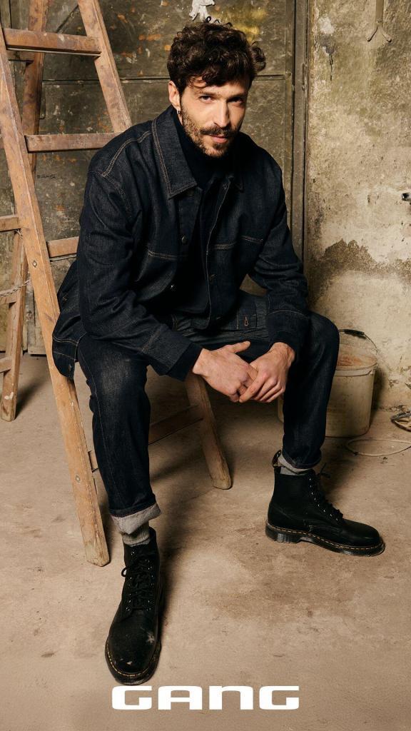 Ein Mann mit lockigem Haar und Bart sitzt auf einer Holzleiter in einem rustikalen Raum und trägt eine dunkle Jeansjacke, Jeans und schwarze Stiefel. Am unteren Rand des Bildes ist das Wort GANG zu lesen.