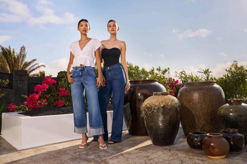 Zwei Frauen posieren im Freien zwischen großen Tongefäßen und Blumen. Die eine trägt eine weiße Bluse, Jeans mit weitem Bein und Sandalen, die andere ein trägerloses schwarzes Oberteil, Jeans mit hoher Taille und Absätze. Heller, sonniger Himmel im Hintergrund. Zwei Frauen posieren im Freien zwischen großen Tongefäßen und Blumen. Die eine trägt eine weiße Bluse, Jeans mit weitem Bein und Sandalen, die andere ein trägerloses schwarzes Oberteil, Jeans mit hoher Taille und Absätze. Heller, sonniger Himmel im Hintergrund.
